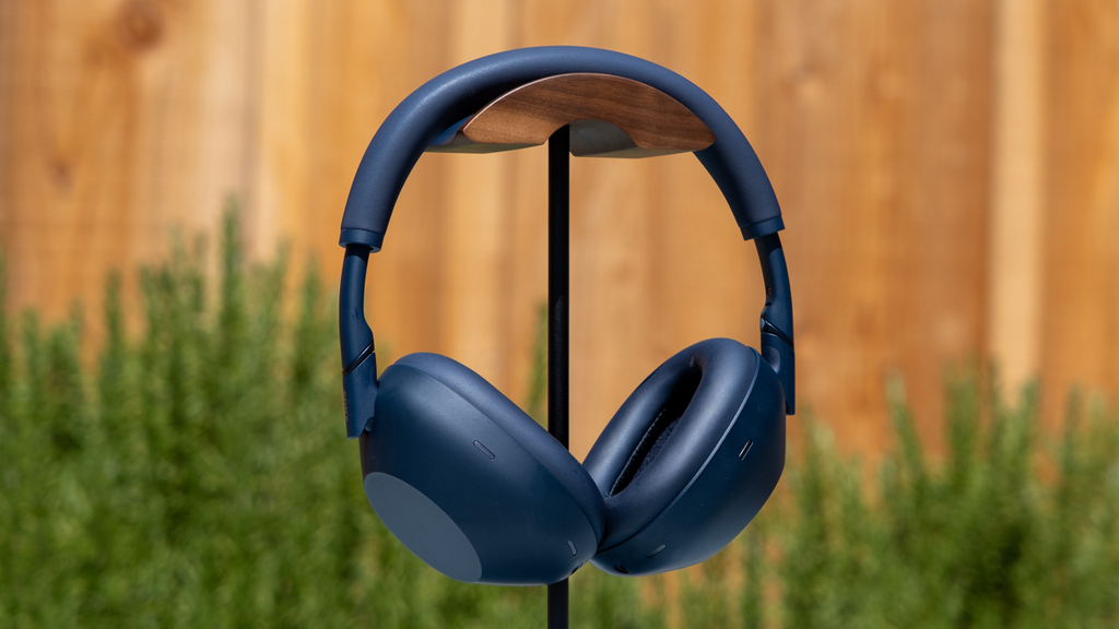 Over-ear navy wireless headphones hanging on a stand against a blurred outdoor background.
