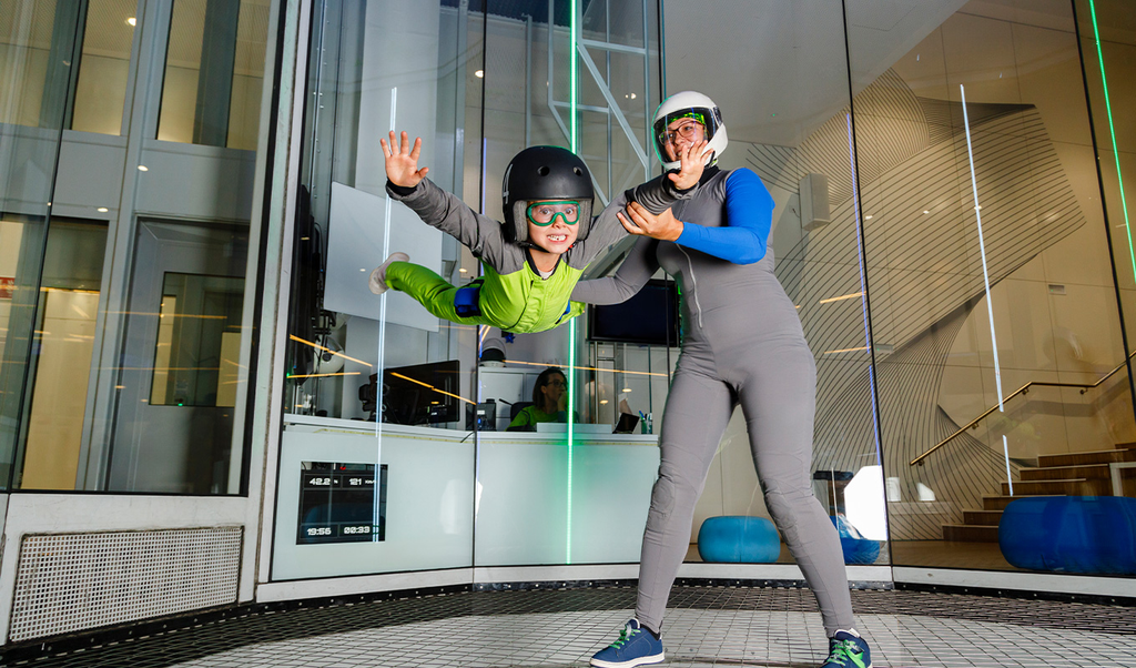 Experience-based gifts. Two people in helmets and safety gear wave and pose in a glass-walled trampoline park